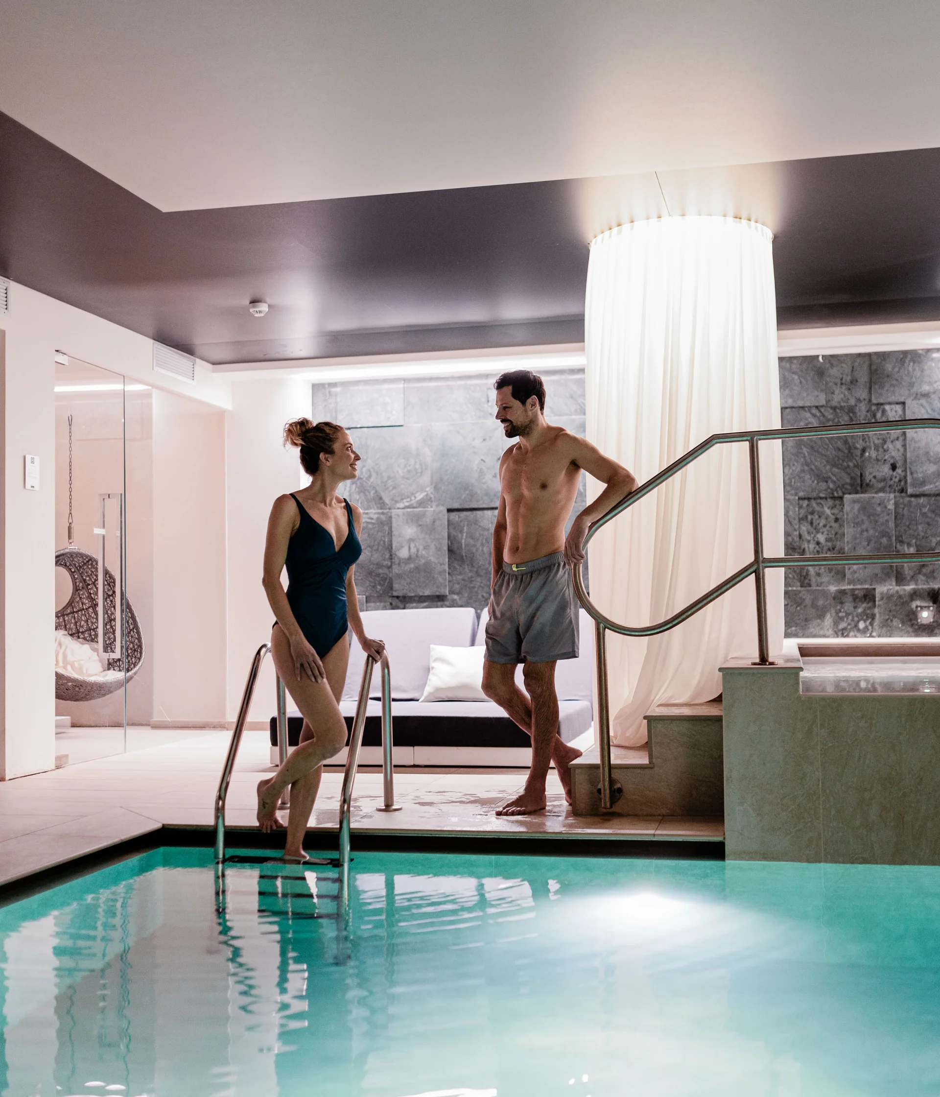 Elgus: hotel a San Giacomo, a Brentonico Coppia che parla vicino a una piscina interna in un ambiente moderno e rilassante